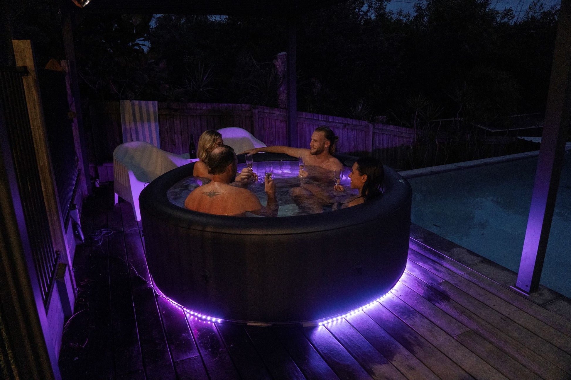 People in a hot tub with purple LED lights on a wooden deck at night.