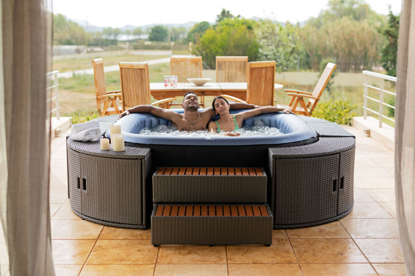 MSpa Two people in a hot tub on a patio with outdoor furniture and greenery.  - Luxe Tub Source