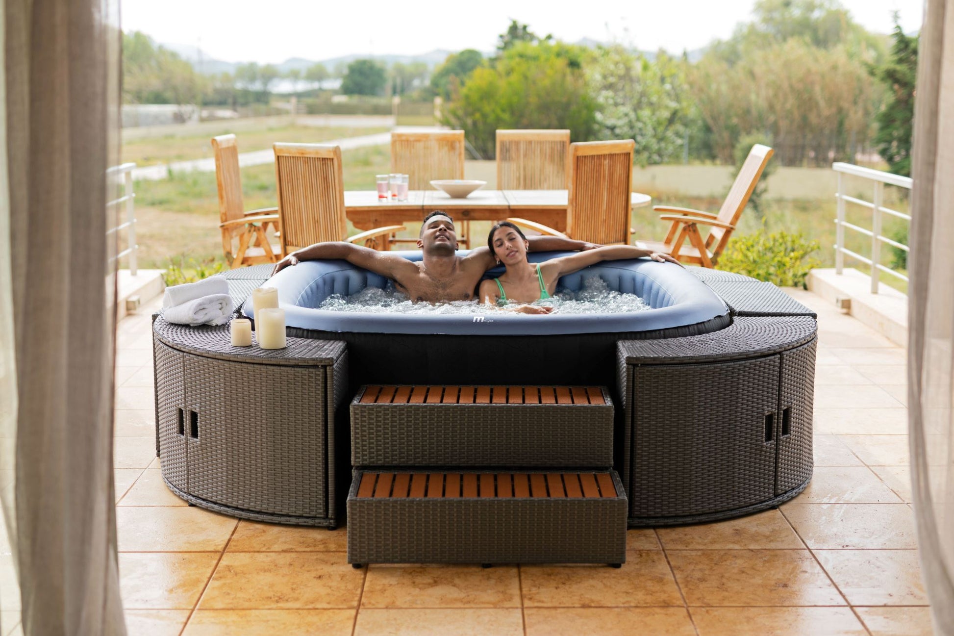 MSpa Two people in a hot tub on a patio with outdoor furniture and greenery.  - Luxe Tub Source
