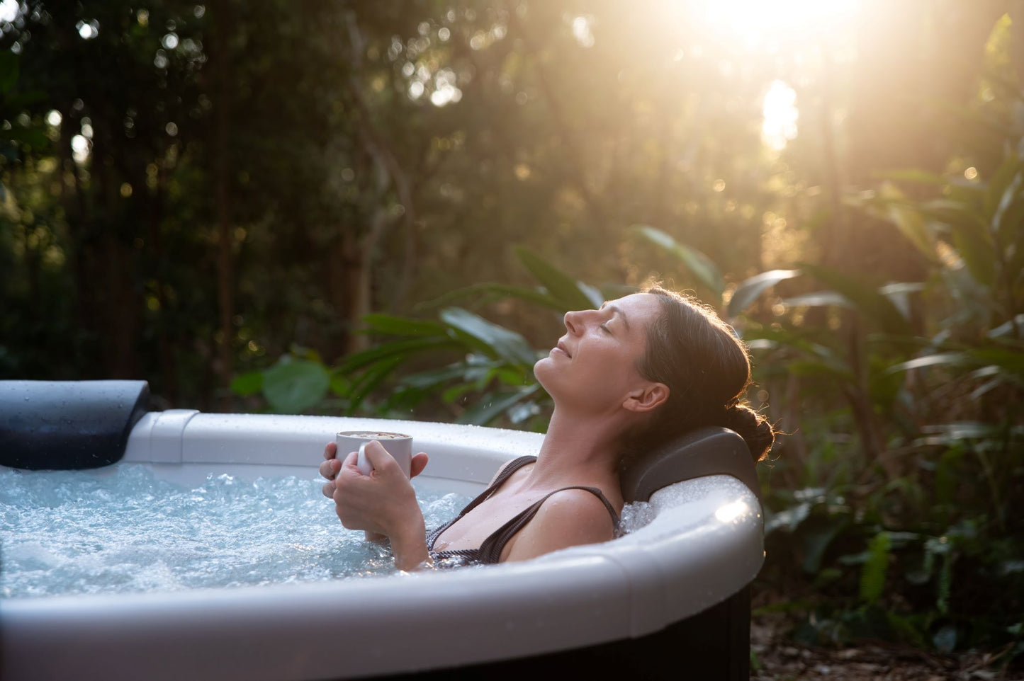 Woman relaxing in a hot tub surrounded by nature with sunlight filtering through the trees.