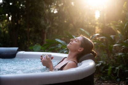 Woman relaxing in a hot tub surrounded by nature with sunlight filtering through the trees.