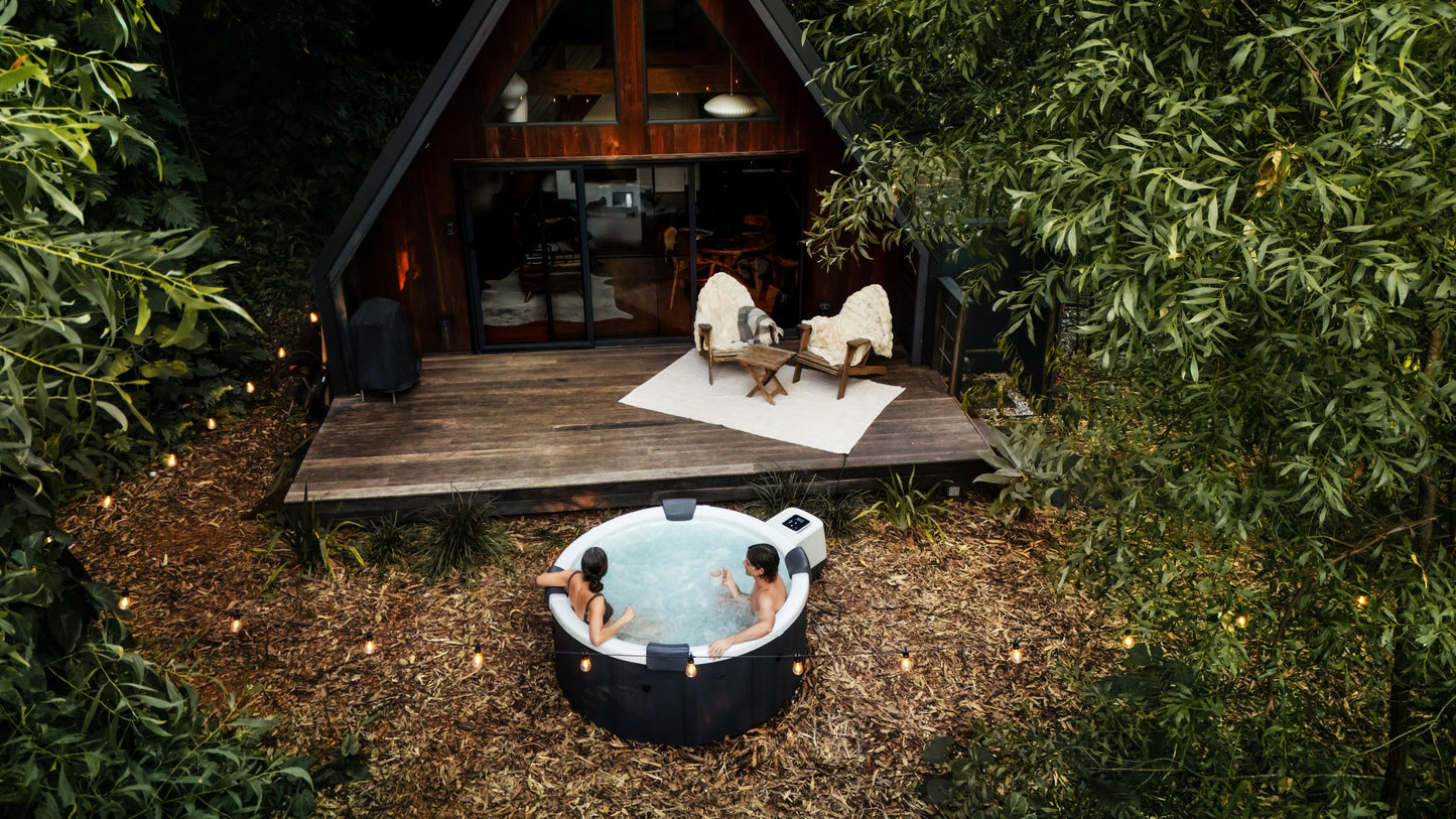 Two people in a hot tub outside a wooden cabin in the woods.