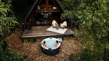 Two people in a hot tub outside a wooden cabin in the woods.